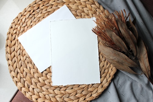 A woven placemat made of natural fibers is laid out with two sheets of plain white paper on top. To the right, a bunch of dried leaves or foliage is placed. The scene is set on a textured, neutral-colored fabric.