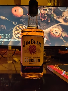 A bottle of Jim Beam Kentucky Straight Bourbon Whiskey is prominently displayed on a desk. In the background, a computer monitor shows a decorative screen with autumn-themed items like pumpkins, leaves, and spices. There are also some desk items such as a small bottle, a closed book or notebook, and what appears to be a mouse pad.