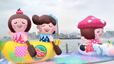 Large, colorful inflatable figures resembling cartoon characters are displayed outdoors near a waterfront. The inflatables include a character sitting on a banana and another with a large toadstool cap. The background features a city skyline with tall buildings and a cloudy sky.