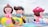 Large, colorful inflatable figures resembling cartoon characters are displayed outdoors near a waterfront. The inflatables include a character sitting on a banana and another with a large toadstool cap. The background features a city skyline with tall buildings and a cloudy sky.