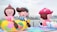 Large, colorful inflatable figures resembling cartoon characters are displayed outdoors near a waterfront. The inflatables include a character sitting on a banana and another with a large toadstool cap. The background features a city skyline with tall buildings and a cloudy sky.