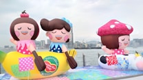 Large, colorful inflatable figures resembling cartoon characters are displayed outdoors near a waterfront. The inflatables include a character sitting on a banana and another with a large toadstool cap. The background features a city skyline with tall buildings and a cloudy sky.