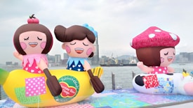 Large, colorful inflatable figures resembling cartoon characters are displayed outdoors near a waterfront. The inflatables include a character sitting on a banana and another with a large toadstool cap. The background features a city skyline with tall buildings and a cloudy sky.