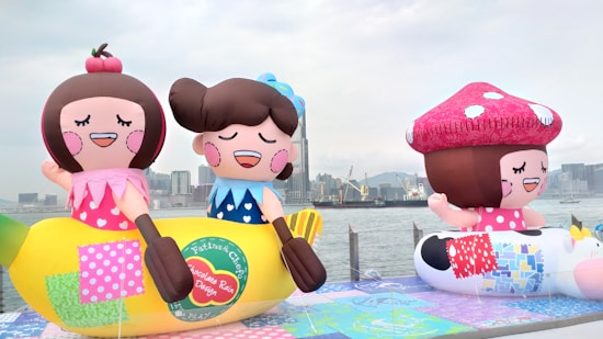 Large, colorful inflatable figures resembling cartoon characters are displayed outdoors near a waterfront. The inflatables include a character sitting on a banana and another with a large toadstool cap. The background features a city skyline with tall buildings and a cloudy sky.
