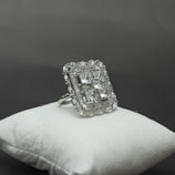 A large, elegant silver ring decorated with multiple square-cut gemstones is displayed on a cushioned white surface against a grey background. The ring's intricate design includes a detailed border surrounding the central stones, giving it a luxurious appearance.