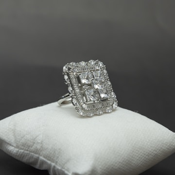 A large, elegant silver ring decorated with multiple square-cut gemstones is displayed on a cushioned white surface against a grey background. The ring's intricate design includes a detailed border surrounding the central stones, giving it a luxurious appearance.