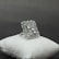 A large, elegant silver ring decorated with multiple square-cut gemstones is displayed on a cushioned white surface against a grey background. The ring's intricate design includes a detailed border surrounding the central stones, giving it a luxurious appearance.