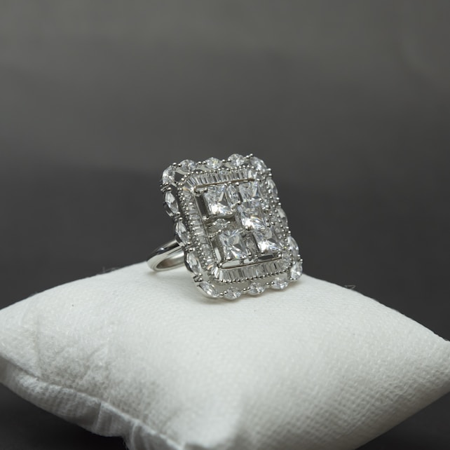 A large, elegant silver ring decorated with multiple square-cut gemstones is displayed on a cushioned white surface against a grey background. The ring's intricate design includes a detailed border surrounding the central stones, giving it a luxurious appearance.