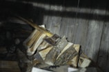A pile of chopped firewood stacked against a dark wooden wall, with an axe placed on top of the logs.