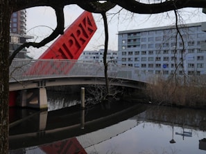 A modern architectural scene featuring a red tilted structure with letters on it, situated next to a bridge with white railings. In the background, there is a large residential building with multiple windows. The image includes tree branches in the foreground and a calm body of water reflecting the scene.