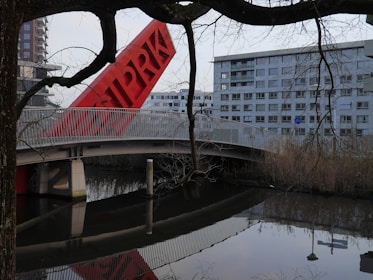 A modern architectural scene featuring a red tilted structure with letters on it, situated next to a bridge with white railings. In the background, there is a large residential building with multiple windows. The image includes tree branches in the foreground and a calm body of water reflecting the scene.