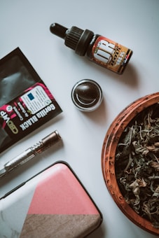 A collection of cannabis-related items arranged on a flat surface. Included are a dropper bottle labeled 'CBD 3%', a small wooden bowl filled with dried herbs, a metallic grinder, rolling papers, a lighter, and a multicolored rectangular case. The items are organized in a casual layout, suggesting a relaxed setting.
