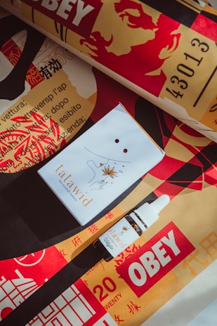 A box and a dropper bottle labelled 'tatawid' are set against a vibrant background featuring the word 'OBEY' along with bold red, yellow, and black design elements and some text in Italian and Chinese. The image features a face in a stencil art style and bold textures.