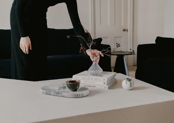 A person dressed in black is arranging a small glass vase with branches on a minimalistic coffee table. The table also holds a stack of books and a round white ceramic piece. In the background, there is a dark sofa and a closed white door. The setting is calm and elegantly styled.