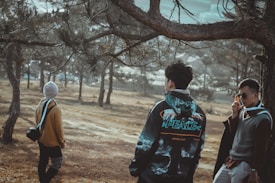 A group of three people is standing in a wooded area. One person is wearing a jacket with the word 'PEACE' visible, another has a backpack and a beanie, and the third is adjusting sunglasses while wearing a sweater. They are surrounded by trees and there is a sense of calm in the natural setting.