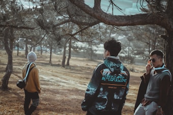 A group of three people is standing in a wooded area. One person is wearing a jacket with the word 'PEACE' visible, another has a backpack and a beanie, and the third is adjusting sunglasses while wearing a sweater. They are surrounded by trees and there is a sense of calm in the natural setting.