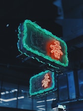 A neon sign with glowing red and turquoise colors is attached to a building at night. The sign features intricate design elements and is mirrored by a similar sign beneath it, creating a visually striking effect in the dimly lit environment.