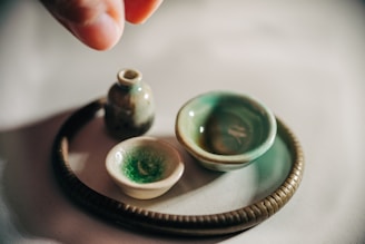 A collection of small, handcrafted pottery items on a smooth surface, including a tiny jug, two small bowls, and a round, textured base. One hand is visible, poised above the arrangement, suggesting careful placement or admiration. The pottery pieces have a rustic appearance with a glaze of greens and browns.
