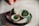 A collection of small, handcrafted pottery items on a smooth surface, including a tiny jug, two small bowls, and a round, textured base. One hand is visible, poised above the arrangement, suggesting careful placement or admiration. The pottery pieces have a rustic appearance with a glaze of greens and browns.
