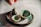 A collection of small, handcrafted pottery items on a smooth surface, including a tiny jug, two small bowls, and a round, textured base. One hand is visible, poised above the arrangement, suggesting careful placement or admiration. The pottery pieces have a rustic appearance with a glaze of greens and browns.