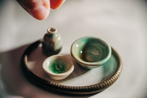 A collection of small, handcrafted pottery items on a smooth surface, including a tiny jug, two small bowls, and a round, textured base. One hand is visible, poised above the arrangement, suggesting careful placement or admiration. The pottery pieces have a rustic appearance with a glaze of greens and browns.