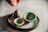 A collection of small, handcrafted pottery items on a smooth surface, including a tiny jug, two small bowls, and a round, textured base. One hand is visible, poised above the arrangement, suggesting careful placement or admiration. The pottery pieces have a rustic appearance with a glaze of greens and browns.