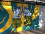 A vibrant mural depicting human lungs with a landscape inside them, representing trees and a blue sky as the inner structures. The surrounding area is painted with abstract patterns in yellow and teal. The mural is located on a paved walkway with dappled sunlight creating shadows.
