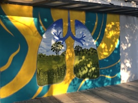 A vibrant mural depicting human lungs with a landscape inside them, representing trees and a blue sky as the inner structures. The surrounding area is painted with abstract patterns in yellow and teal. The mural is located on a paved walkway with dappled sunlight creating shadows.