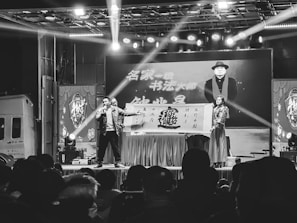 A stage setup with a performer holding a microphone and another person holding a large sign. Bright stage lights illuminate the area, and there is a crowd watching the performance. A large screen in the background displays an image of a man wearing a hat and scarf with text beside him.