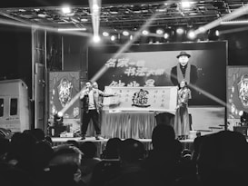 A stage setup with a performer holding a microphone and another person holding a large sign. Bright stage lights illuminate the area, and there is a crowd watching the performance. A large screen in the background displays an image of a man wearing a hat and scarf with text beside him.