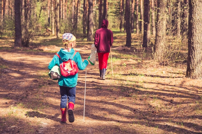 A child wearing a colorful cap, blue jacket, red backpack, and rubber boots is walking through a forest trail. The child is holding hiking poles and a soccer ball under one arm. Another person, wearing a red hoodie and pants, is walking ahead on the same path. The background is filled with tall trees and sunlight filtering through leaves, creating dappled shadows on the ground.