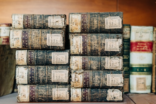 A stack of old, worn-out books with leather bindings and visible labels. The spines show signs of age, with some discoloration and frayed edges. These books are organized in two stacks on a wooden surface, with additional books blurred in the background.