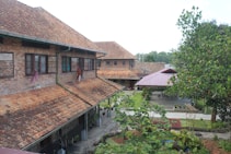A rustic brick building with sloped, tiled roofs surrounded by lush greenery. Windows are open, and clothing hangs outside to dry. A red-roofed shelter and garden area with various plants can be seen nearby.