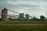 An industrial facility with multiple large, connected structures and conveyor belts spread across a grassy area under an overcast sky.