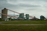 An industrial facility with multiple large, connected structures and conveyor belts spread across a grassy area under an overcast sky.