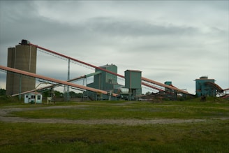 An industrial facility with multiple large, connected structures and conveyor belts spread across a grassy area under an overcast sky.