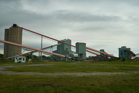 An industrial facility with multiple large, connected structures and conveyor belts spread across a grassy area under an overcast sky.