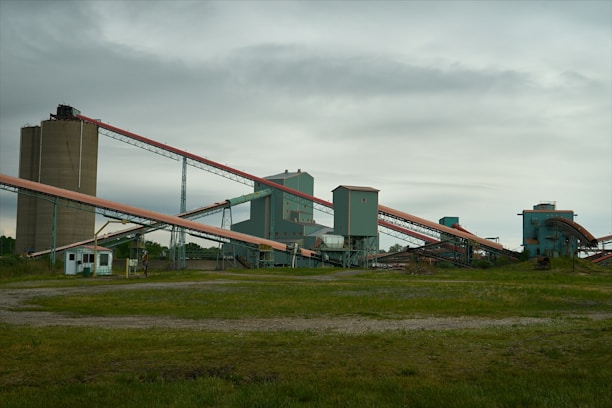 An industrial facility with multiple large, connected structures and conveyor belts spread across a grassy area under an overcast sky.
