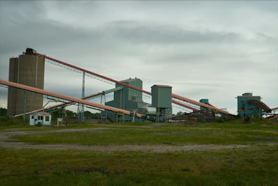 An industrial facility with multiple large, connected structures and conveyor belts spread across a grassy area under an overcast sky.