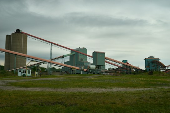 An industrial facility with multiple large, connected structures and conveyor belts spread across a grassy area under an overcast sky.