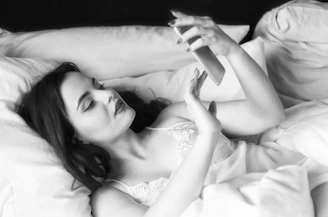 A woman with dark hair is lying in bed, looking at her smartphone. She is wearing a lace-trimmed top and appears to be relaxed, with a pillow behind her head. The image is in black and white, creating a soft, calm atmosphere.