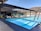 An outdoor pool with clear blue water is surrounded by modern architecture. Several people are seated at tables under a shaded patio area, and there are trees and grassy hills in the background under a bright, clear sky.