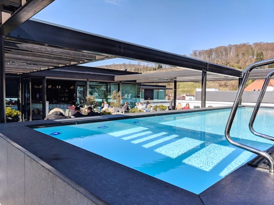 An outdoor pool with clear blue water is surrounded by modern architecture. Several people are seated at tables under a shaded patio area, and there are trees and grassy hills in the background under a bright, clear sky.