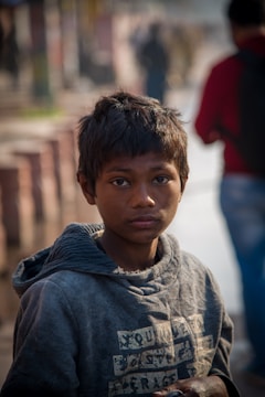 A young boy with a serious expression stands in a city setting. He is wearing a worn grey hoodie, and his hair appears slightly unkempt. The background is blurred with people walking, capturing a busy urban atmosphere.