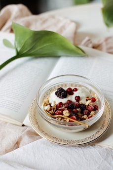 A bowl of yogurt topped with granola, blackberries, and red berries placed on a decorative plate over an open book. A large green leaf and soft beige fabric are in the background, enhancing the cozy and inviting atmosphere.