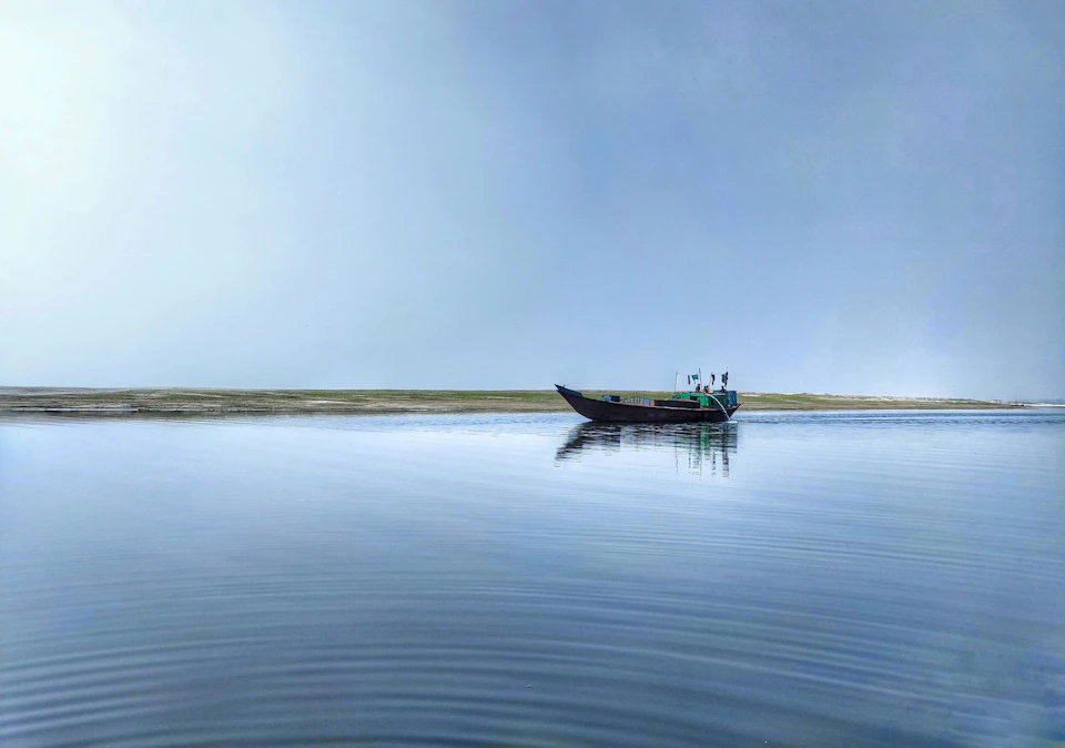 The image depicts a serene scene on a body of water, with two individuals aboard a small boat. The boat is painted black and contrasts against the light blue-green color of the water. One person is seated at the front of the boat, while the other stands near the back. Both individuals are wearing green shirts that match the color of the water. In the background, there's an island visible on the right side of the image.