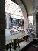 A view of a traditional market scene with stone arches framing the entrance to a covered section. Inside, there are stalls displaying various goods such as shoes and clothes. Several small tables and plastic chairs are scattered, giving a casual and inviting atmosphere. Turkish flags hang from the ceiling, and the stone architecture suggests an old building.