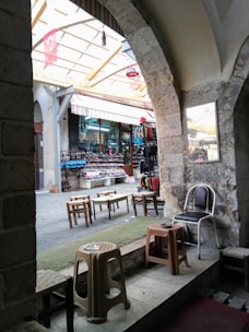 A view of a traditional market scene with stone arches framing the entrance to a covered section. Inside, there are stalls displaying various goods such as shoes and clothes. Several small tables and plastic chairs are scattered, giving a casual and inviting atmosphere. Turkish flags hang from the ceiling, and the stone architecture suggests an old building.