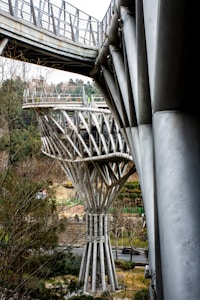 A modern architectural structure composed of large metal beams and pillars, resembling a bridge or viewing platform in a park setting. The design features intricate, vertical, and diagonal supports, with trees and roads visible in the background, suggesting an integration with nature.