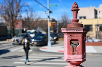 An old-fashioned red fire alarm box with the words 'FDNY' and instructions visible on it is mounted on a metal post. The background shows a street scene with cars at an intersection and a pedestrian crossing the road. Buildings, trees, and street signs are present, and the sky is clear with a few clouds.